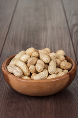 Close-up photo of the wooden bowl full of roasted unshelled peanuts on the wooden table background with copy space.