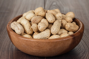 Close-up photo of the wooden bowl full of roasted peanuts on the wooden table background.