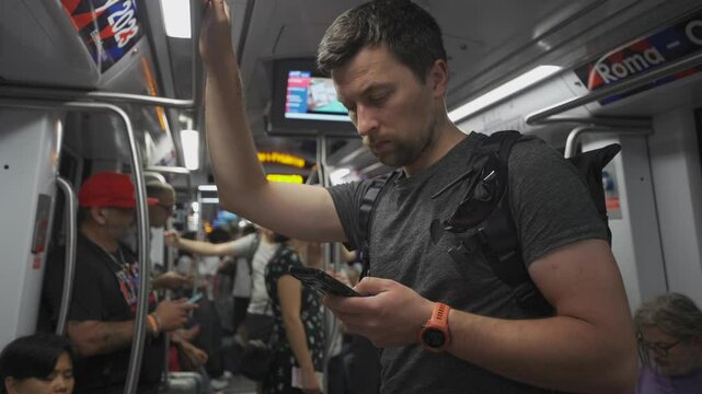 Male passenger on Roman subway in Italy uses smartphone in crowded metro train in summer. Male using phone while traveling in full train car of underground in Rome. Theme public transportation in Rome