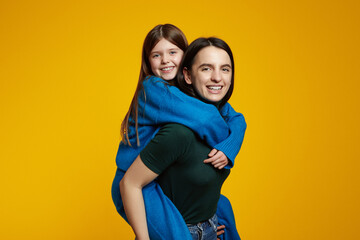 Cheerful smiling young woman and little kid girl sisters wearing comfy sweaters hugging and looking at camera, isolated over yellow background