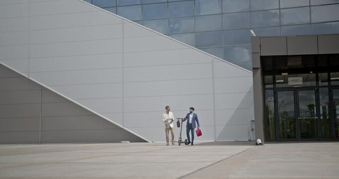 Two businessmen walk and talk outside a corporate building. One holds an electric scooter. They carry bags and documents.