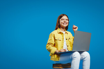 Enthusiastic youngster wearing glasses and a yellow jacket works on a laptop with a big smile,...