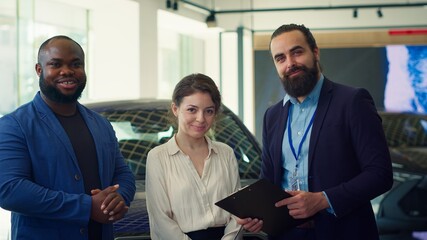 Portrait of cheerful salesman talking with customers, showcasing dealership vehicles and explaining...