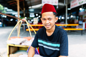 Portrait of young indonesian street vendor in songkok hat selling local food at street food stall...