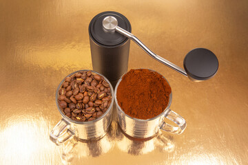 A manual coffee grinder sits between two glass mugs, one with roasted coffee beans, the other with...