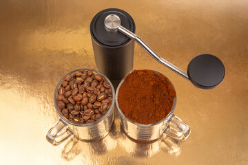 A manual coffee grinder sits between two glass mugs, one with roasted coffee beans, the other with...