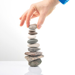 A hand carefully places a stone on a balanced stack of smooth pebbles on a reflective white surface against a white background. Ideal for themes of balance, mindfulness, and zen.