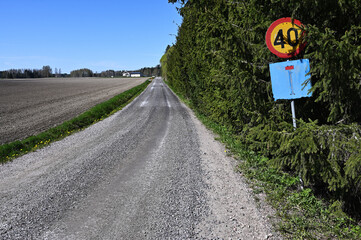 a tranquil rural gravel road running between a freshly plowed field and a dense line of coniferous...