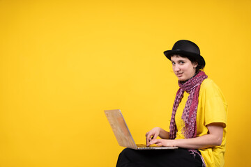 Young artist using laptop wearing bowler hat and colorful scarf on yellow background