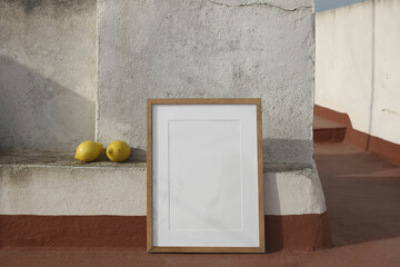Blank wooden picture frame leaning against textured wall on Spanish house rooftop. Two yellow lemons on a concrete ledge. Natural sunlight, rustic setting, calm mood. Minimalist summer outdoor scene.