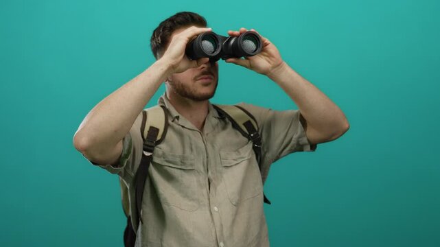 Young man with beard wearing backpack and looking through binoculars against isolated green background in casual tourist exploration scene.