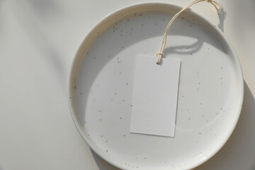 Minimalist flat lay. Blank paper gift tag, label with string on a white speckled ceramic plate....
