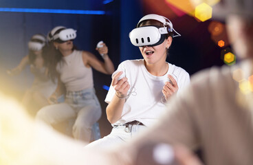 Young woman sitting on stool and enjoying virtual reality game with VR headset and controllers in...