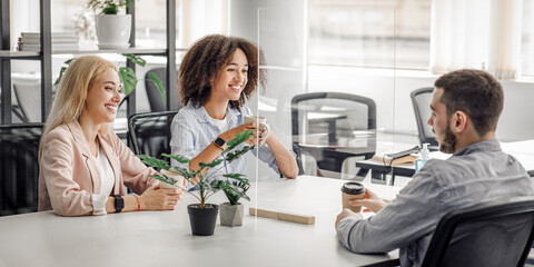 Modern coffee break in restroom during covid epidemic, new normal and health protection. Multiracial colleagues with cups smiling and talking through protective glass in interior, empty space