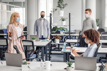 Obraz premium Meeting in office and instructions from boss during social distancing and virus outbreak. Young business woman in protective mask in suit with tablet in hand speaks to workers at workplace