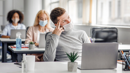 Creative ideas and brainstorming during work and social distancing. Guy in protective mask thinks and looks out the window, sitting at table with laptop with antiseptic in office interior with