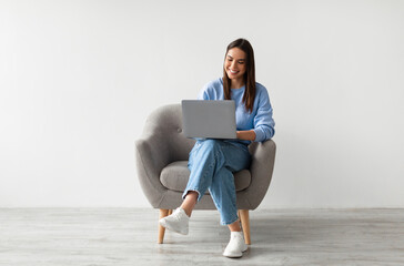 Fototapeta na wymiar Happy Caucasian lady sitting in armchair with modern laptop, studying remotely or having business meeting against white studio wall, full length. Young woman having online class or watching webinar