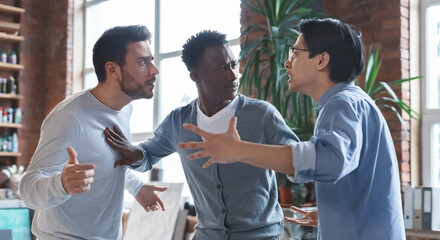 Office conflict. Angry multiracial young men fighting at workplace, afro guy standing between them
