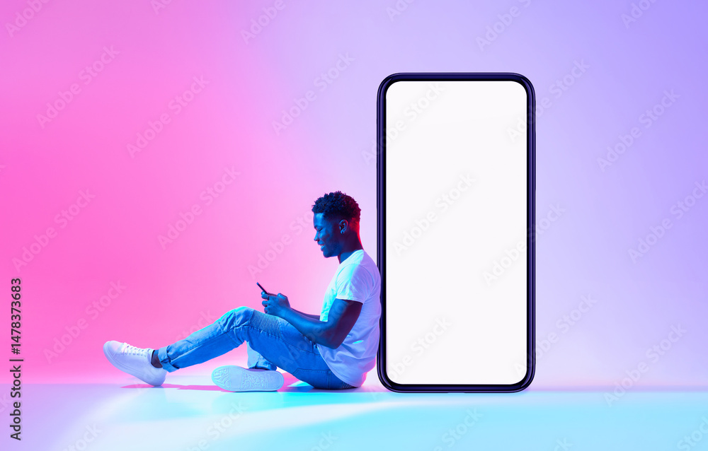 Poster Young black man sitting next to huge smartphone with empty screen, advertising new mobile application or website, using modern gadget in neon light. Cellphone display mockup