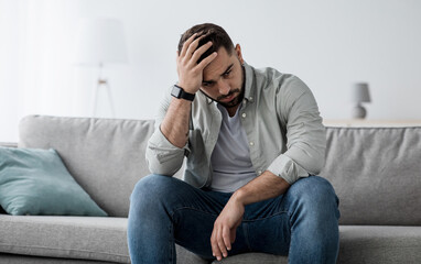 Plakat Unhappy man touching hair, sitting on sofa at home, thinking about problems. Upset guy feeling lonely, sad, have psychological and mental troubles, suffering from bad relationship or break up