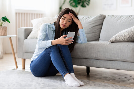 Smartphone Addiction And Depression. Upset Asian Girl Sitting On Floor At Home With Smartphone, Looking Sadly To Device Screen