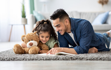 Loving arabic dad reading book to his cute little daughter at home, happy middle eastern father and...