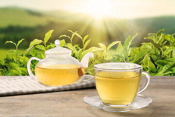 Green tea in cup and teapot on wooden table against tea plantation in mountains
