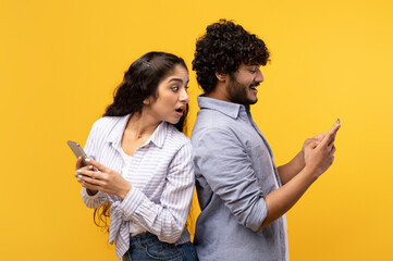 Shocked indian woman spying on her smiling boyfriend who using smartphone, chatting or scrolling...