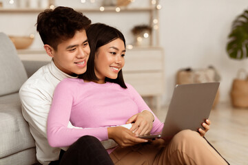 Fototapeta premium Happy Korean Couple Using Laptop Watching Movie Online Enjoying Weekend, Relaxing Sitting On Floor At Home. Evening Together, Stay-At-Home Date Leisure Concept