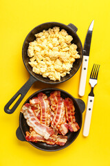 Frying pans of tasty scrambled eggs and bacon on yellow background
