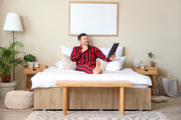 Young happy man in checkered pajamas with magazine lying on bed at home