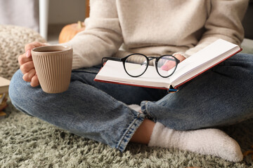 Woman with book, eyeglasses and coffee cup sitting on carpet at home, closeup