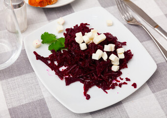 On table there is plate with portion of dietary and healthy food - salad of stewed beet with cubes of cheese, decorated with peach of coriander.
