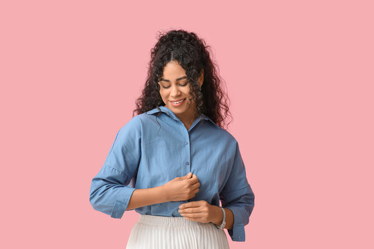 Pretty young African-American woman buttoning up stylish blue shirt on pink background - Powered by Adobe
