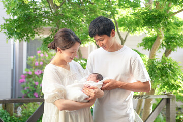A Japanese mother and father in their twenties hold their 7-day-old newborn baby on a wooden deck with spring greenery. This image shows the love shared by new parents in a peaceful outdoor setting.