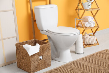 Interior of restroom with toilet bowl, basket with towel and shelving unit near yellow wall