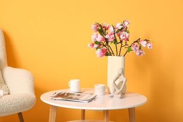 Armchair and coffee table with blooming magnolia branches near yellow wall in room
