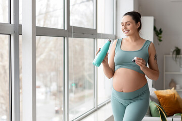 Sporty pregnant woman with water bottle near window at home