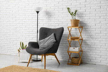 Comfortable black armchair with wooden shelving unit and floor lamp near white brick wall in room