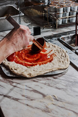 Chef spreads tomato sauce on fresh pizza dough with a ladle in professional pizzeria kitchen. Artisanal food preparation on marble countertop showcases authentic Italian culinary craftsmanship.