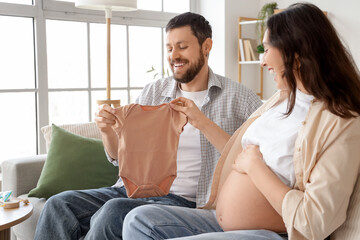 Happy pregnant couple with baby bodysuit sitting on sofa at home