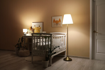 Interior of children's bedroom with baby crib, table and glowing lamps near beige wall at night
