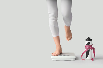 Legs of sporty woman measuring her weight on scales and water bottle against light background