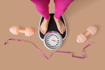 Female legs on weight scales with dumbbells and tape measure against beige background, top view