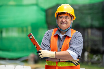 Fototapeta premium Friendly mature construction worker man wea yellow safety helmet sitting on brick wall outdoors smiling at camera in a green park with trees and bushes on a bright day