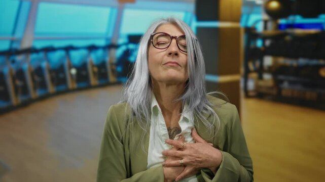 Senior woman with grey hair in gym placing hands on chest indicating discomfort against a backdrop of exercise equipment and large windows.