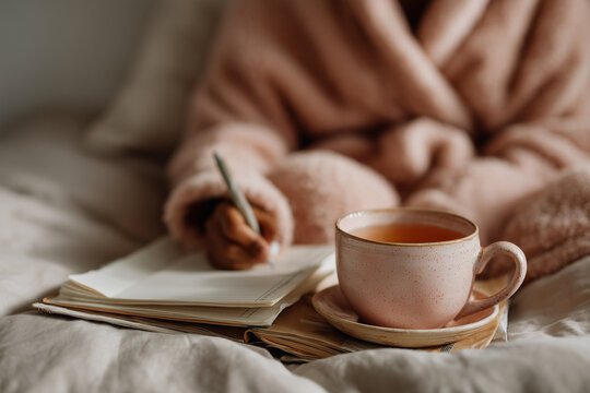 Woman enjoying a quiet morning in bed, writing in her personal journal while sipping a warm cup of tea, creating a cozy and relaxing atmosphere