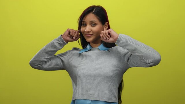 Woman covering ears with fingers against yellow background offering a playful expression in isolated setting.