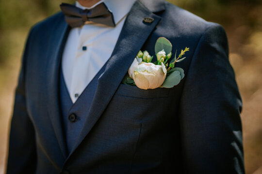 Close-up of a groom in a navy blue suit with a white rose boutonniere and a dark bow tie, dressed for a wedding ceremony outdoors..
