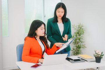 Two Asian business woman are discussing business reports and analyzing data at a desk. One is using...